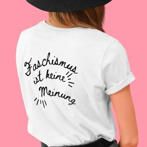 May include: White t-shirt with the German text "Faschismus ist keine Meinung" in black script. The shirt has short sleeves and a rolled hem. The background is a solid pink color.