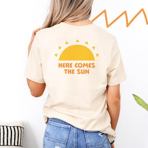 Op de afbeelding: Een licht beige t-shirt met een gele zon graphic en de tekst "Here Comes The Sun" gedrukt op de achterkant.
