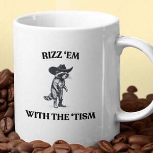 May include: White ceramic coffee mug with the text "RIZZ 'EM WITH THE 'TISM" and a cartoon raccoon wearing a cowboy hat. The mug is surrounded by coffee beans, with steam rising from the mug.