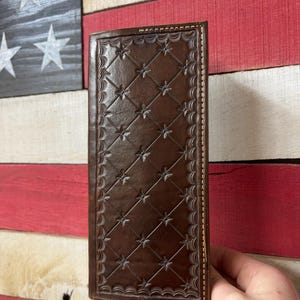 May include: A brown leather wallet with a diamond and star pattern. The wallet is held in a hand and has a decorative border. The background features a wooden American flag.