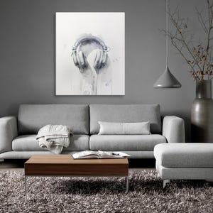 May include: A gray sofa with a throw blanket and a wooden coffee table in front of it. A gray ottoman is to the right of the sofa. A gray wall with a white canvas print of a pair of headphones hanging on it.