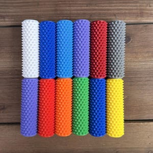 May include: A collection of twelve colorful, cylindrical massage rollers with textured surfaces. The rollers are arranged in two rows of six, showcasing a variety of colors including white, blue, purple, red, orange, green, and yellow. Each roller is approximately 4 inches in length.