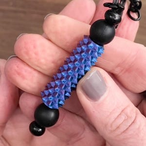 Peut inclure: Gros plan sur un bracelet de perles bleu et violet avec des perles noires et un centre cylindrique texturé. Le bracelet est tenu dans une main avec un ongle peint.