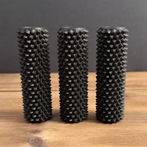 Op de afbeelding: Drie cilindrische, donkergrijze objecten met een getextureerd, stekelig oppervlak. De objecten staan rechtop op een houten oppervlak. De achtergrond is effen donkergrijs. Elk object is ongeveer 10 cm hoog.