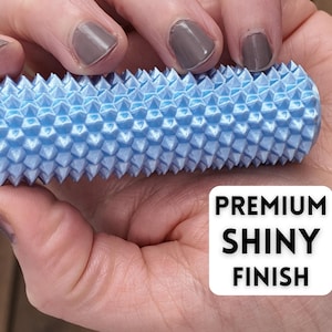 K&ouml;nnte beinhalten: Ein hellblaues, zylindrisches Massageger&auml;t mit einer strukturierten, stacheligen Oberfl&auml;che. Das Werkzeug wird in einer Hand gehalten und zeigt den Text "PREMIUM SHINY FINISH" in einem wei&szlig;en Feld.