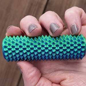 May include: A blue and green massage roller with a textured surface. The roller is held in a hand and has a series of raised, pointed protrusions. The color scheme is a gradient from blue to green.