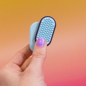 May include: A light blue and navy blue oval-shaped beauty tool with a textured surface. The tool is held against a light blue oval-shaped piece. The background is a gradient of orange and pink.
