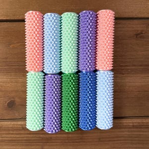 Op de afbeelding: Een verzameling van tien kleurrijke schuimrollers in verschillende tinten roze, blauw, groen en paars. Elke roller heeft een textuuroppervlak met kleine, gelijkmatig verdeelde uitsteeksels. De rollers zijn gerangschikt in een rechthoekig patroon op een houten oppervlak.