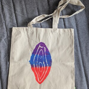 Könnte beinhalten: Beige Canvas-Tragetasche mit farbenfrohem, abstraktem Design. Das Design zeigt eine stilisierte, geschichtete Form in Lila, Blau und Rot. Die Tasche hat zwei lange, beige Griffe zum Tragen.