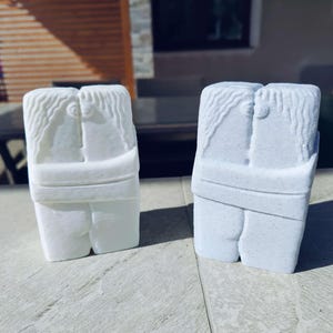 Può includere: Due sculture bianche di figure che si abbracciano. Le figure sono scolpite da un materiale solido, con dettagli come capelli e vestiti. Le sculture sono poste su una superficie chiara, con uno sfondo sfocato all'aperto.