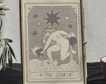 May include: A framed print of the Star tarot card, depicting a nude figure and stars. A small vase with foliage and a small bowl with a spoon are on a dark surface. The image has a neutral colour palette.