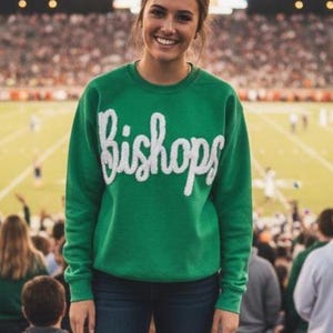 May include: A green sweatshirt with the word "Bishops" in white script. The sweatshirt is being worn at a football game. The person is wearing blue jeans and is smiling at the camera.