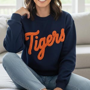 Puede incluir: Sudadera azul marino con la palabra "Tigers" en escritura naranja. Se usa con vaqueros azul claro y zapatillas blancas. La persona está sentada en un sofá gris claro en una habitación luminosa.