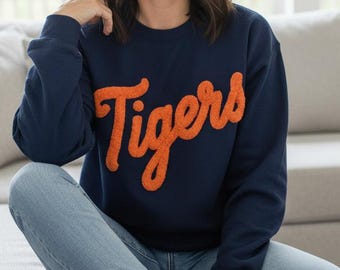 Puede incluir: Sudadera azul marino con la palabra "Tigers" en escritura naranja. Se usa con vaqueros azul claro y zapatillas blancas. La persona está sentada en un sofá gris claro en una habitación luminosa.