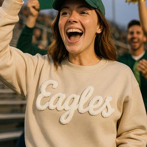 Puede incluir: Una sudadera beige con la palabra "Eagles" en letras blancas y esponjosas. Una persona lleva la sudadera y una gorra verde, haciendo el signo de la victoria. Un cartel que dice "GO GAL" está en el fondo.