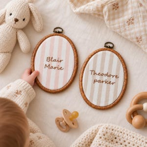 May include: Two oval wall plaques with dark wood frames and metal hangers. One plaque features pink and white vertical stripes with the name Blair Marie in brown script. The other plaque features beige and white vertical stripes with the name Theodore parker in brown script.