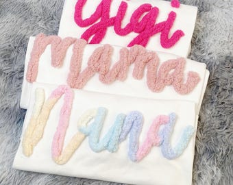 May include: Three white t-shirts with colorful, textured lettering. The top shirt has "Gigi" in bright pink. The middle shirt says "Mama" in a soft pink. The bottom shirt says "Nana" in a pastel rainbow of colors.