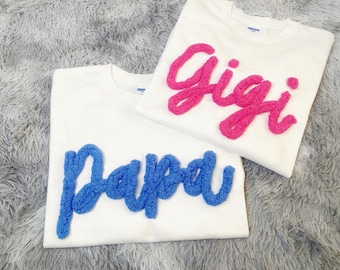 May include: Two white t-shirts with text. One shirt has the word "Gigi" in pink, textured lettering. The other shirt has the word "Papa" in blue, textured lettering. The shirts are on a gray, fluffy surface.