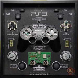 Könnte beinhalten: Eine schwarze und grüne PlayStation 3 Konsole, die zerlegt und in einem gerahmten Kunstwerk ausgestellt ist. Die Komponenten der Konsole sind in Japanisch und Englisch beschriftet, darunter die Gehäusehülle, das Motherboard, der DC-Ladeanschluss, die Vibrationsmotoren und die Analogsticks.