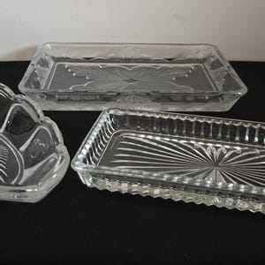 May include: Three clear glass serving dishes. One is a scalloped bowl with a sunburst design. The other two are rectangular trays with etched patterns. The trays have different edge designs.