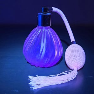 May include: A vintage perfume bottle with a bulbous, ribbed purple glass body and a black cap. It has a white, woven atomizer and tassel, set against a dark background. The bottle is approximately 15 cm tall.