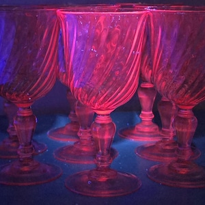 May include: Six pink-tinted stemmed glasses are displayed. Each glass has a spiral pattern on the bowl and an ornate stem. The glasses are grouped closely, giving a sense of fullness. The lighting creates a pink glow, emphasising the glassware.