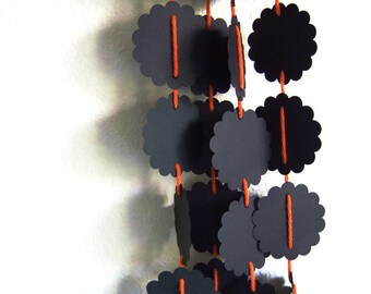 May include: A decorative garland featuring black, scalloped-edge paper shapes strung together with orange twine. The garland hangs vertically against a white wall, creating a striking contrast. The design is simple yet visually appealing.