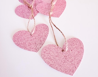 May include: Pink glitter heart-shaped gift tags with twine strings. Several heart tags are visible, some overlapping, against a white background. The tags have a sparkly, textured surface, ideal for Valentine's Day or other celebrations.