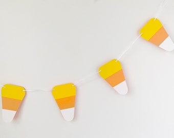 May include: A string of paper candy corn cutouts in yellow, orange, and white, strung on white twine. The candy corn cutouts are hanging against a white background.