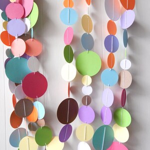 May include: A colorful paper circle garland with various shades of blue, green, red, orange, yellow, and purple. The circles are strung together with white thread and hang from a white wall.