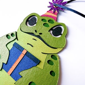May include: A green frog wearing a party hat with a purple flower on top. The frog is holding a blue and orange gift box.