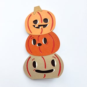May include: A stack of three paper pumpkins with different facial expressions. The top pumpkin has a toothy grin, the middle pumpkin has wide eyes, and the bottom pumpkin has a wide smile.