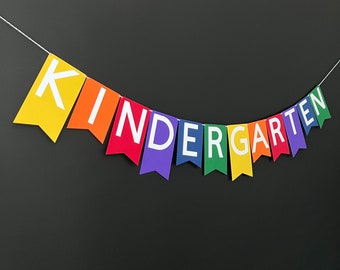 May include: A colorful banner spelling "KINDERGARTEN" hangs against a dark background. Each flag is a different color of the rainbow, with white letters. The banner is strung on a white string, creating a festive and educational decoration.