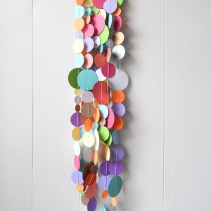 May include: A colorful paper circle garland with a variety of colors, including pink, orange, green, blue, and purple. The garland is hanging from a white wall.