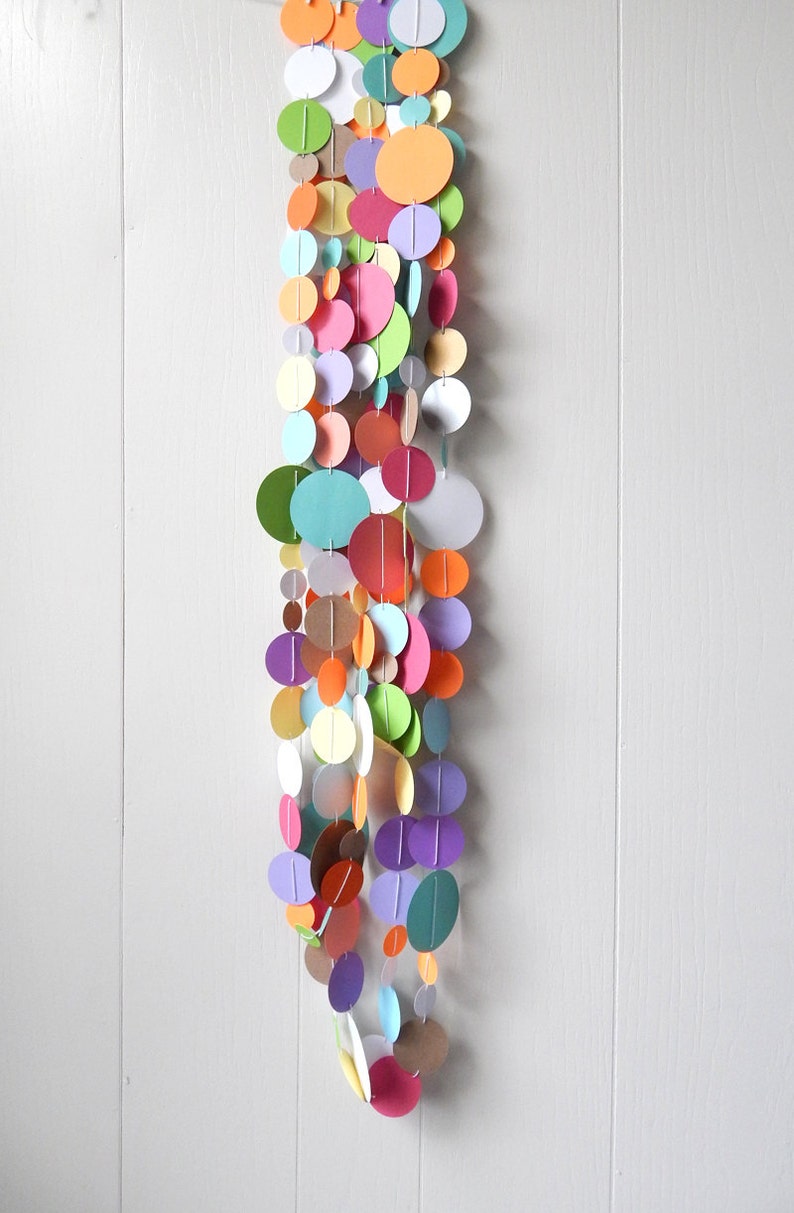 May include: A colorful paper circle garland with a variety of colors, including pink, orange, green, blue, and purple. The garland is hanging from a white wall.