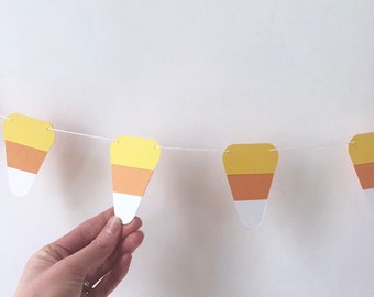 May include: A festive garland featuring candy corn shapes in yellow, orange, and white. The paper cutouts are strung together with white string, perfect for Halloween or autumn decorations. The garland is displayed against a plain white background.