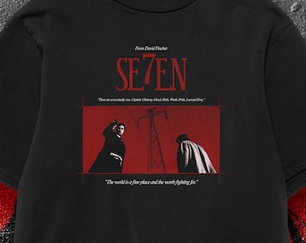 Op de afbeelding: Zwart T-shirt met het woord "SE7EN" in het rood en een rode rechthoekige afbeelding eronder. De afbeelding bevat silhouetten en tekst uit de film. Het shirt heeft een ronde hals en korte mouwen.