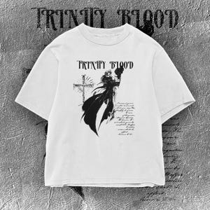 Può includere: Una t-shirt bianca a maniche corte con una stampa grafica nera sul petto. Il design include il testo "TRINITY BLOOD" sopra un'illustrazione di una figura che impugna un'arma accanto a una croce, accompagnata da testo in corsivo.