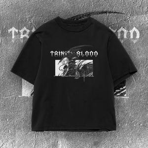 Può includere: Una t-shirt nera a maniche corte con una stampa grafica bianca sul petto. Il design include il testo "TRINITY BLOOD" in un carattere stilizzato sopra un'illustrazione di un personaggio con un uccello, all'interno di una cornice bianca.