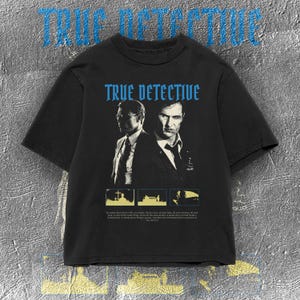 Pode incluir: T-shirt preta com a inscrição "TRUE DETECTIVE" em azul. A camisa apresenta um gráfico de dois homens de terno, com imagens adicionais abaixo. A camiseta tem mangas curtas e gola redonda.