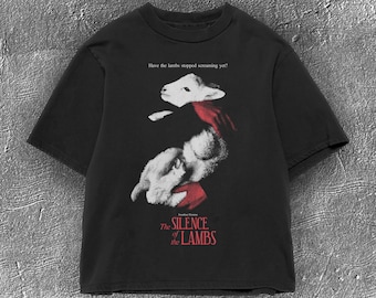 Op de afbeelding: Zwart t-shirt met een afbeelding uit "The Silence of the Lambs". Het ontwerp toont een lammetje met een rode sjaal en de tekst "Have the lambs stopped screaming yet?" en "The Silence of the Lambs."