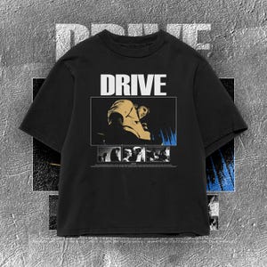 May include: Black t-shirt with the word "DRIVE" in white block letters. The shirt features a graphic of a person in a yellow shirt and a series of black and white images below. The background is a textured gray.