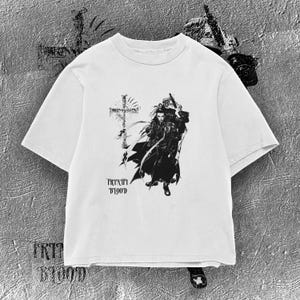 May include: A white short-sleeved t-shirt featuring a black graphic print of a character holding a weapon next to a cross. The text Trinity Blood is printed in a stylised font below the cross. The design is centred on the front of the garment.