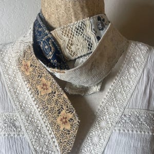 May include: A multi-patterned scarf with floral, paisley, and lace designs. The scarf features a combination of beige, navy, and cream colors. The scarf is wrapped around a mannequin head, showcasing its design and texture.