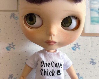 Op de afbeelding: Een pop die een wit T-shirt draagt met de tekst "One Cute Chick" en een kleine vogelafbeelding. De pop heeft paars haar en groene ogen.