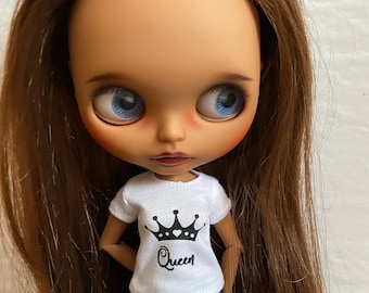Op de afbeelding: Een pop met lang bruin haar die een wit T-shirt draagt met een zwarte kroon en het woord "Queen" erop gedrukt. De pop draagt ook een blauwe jeans.
