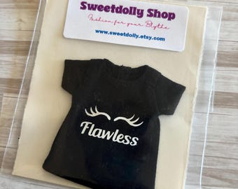 Op de afbeelding: Een zwart t-shirt met het woord "Flawless" in witte letters en witte wimperafbeeldingen. Het shirt is verpakt met een label met de tekst "Sweetdolly Shop Fashion for your Blythe" en het websiteadres.