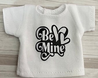 Op de afbeelding: Een klein wit t-shirt met de tekst "Be Mine" in zwart schrift. Het ontwerp bevat een hartvorm. Het shirt heeft korte mouwen en een ronde halslijn.