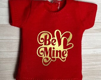 Op de afbeelding: Een klein, rood t-shirt met een gouden folie ontwerp met de tekst "Be Mine" in een decoratief lettertype. Het shirt is gemaakt van een zachte stof en heeft korte mouwen en een ronde halslijn. De tekst staat in het midden op de voorkant.
