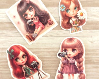 Op de afbeelding: Vier kleurrijke stickers met illustraties van meisjes met grote ogen. Elke sticker heeft een ander ontwerp, waaronder een meisje dat een camera vasthoudt, een meisje dat een bloem vasthoudt en een meisje met een kleinere pop.